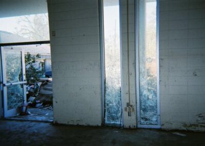 Tall narrow windows inside the Pass Christian Library are cracked and shattered, with debris and damaged vegetation visible outside the open doorway. The interior walls and floors show heavy water staining.