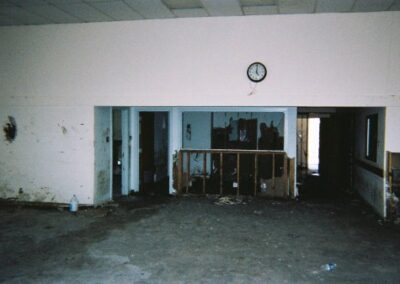 A large interior room of the Pass Christian Library shows mud‑stained walls, exposed lower drywall, and debris scattered across the bare floor. A clock hangs above a damaged doorway with darkened rooms visible beyond.