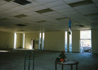 A large empty room inside the Pass Christian Library shows mud‑stained floors, missing furnishings, and sunlight coming through broken windows and an open doorway. A small table with a damaged holiday decoration sits in the foreground.