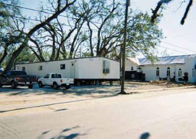 Portable library trailers sit along a roadside beside storm‑damaged buildings and stripped trees in Pass Christian. Vehicles are parked nearby among scattered debris.