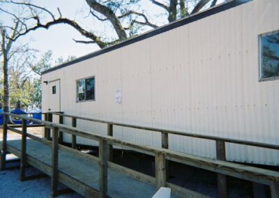A portable trailer used as a temporary Pass Christian Library facility stands on a wooden ramp surrounded by storm‑damaged trees. A small handwritten sign is taped beside one of the windows.