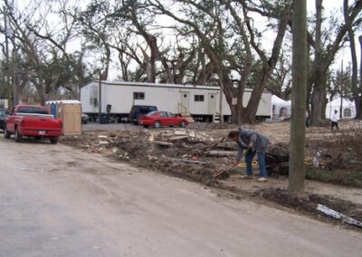 A person clears debris along a roadside lined with portable trailers and damaged trees near the Pass Christian Library site. Cars and scattered storm wreckage fill the area.