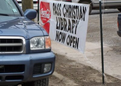 A banner reading “PASS CHRISTIAN LIBRARY NOW OPEN” is attached to metal poles beside a parked vehicle. The sign hangs outdoors near a gravel‑covered area.