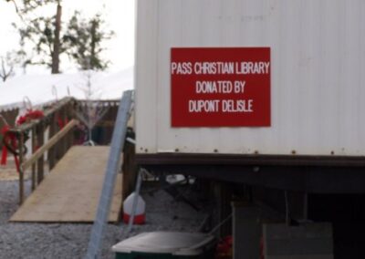 A portable building with a red sign reading “PASS CHRISTIAN LIBRARY DONATED BY DUPONT DELISLE” stands on gravel with a ramp leading to its entrance. Holiday decorations hang along the railings.