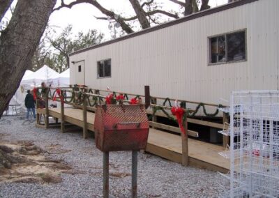 A portable library trailer is decorated with garland and red bows along its wooden ramp. Nearby, display racks and storm‑damaged trees surround the temporary facility.