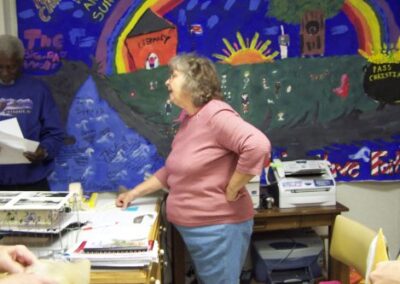 Inside a temporary library room, a bright mural depicting a rainbow, sun, and landscape covers the wall above desks filled with papers and office equipment. People are gathered nearby, appearing to discuss or review materials.