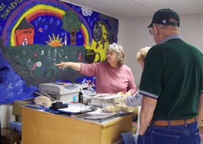 Inside a temporary library space, a colorful mural with a rainbow and the words “We Are Survivors” hangs on the wall behind a desk covered with papers and office supplies. People stand nearby, engaged in conversation.