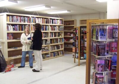 Two people stand talking inside the temporary Pass Christian Library, surrounded by fully stocked bookshelves and a rotating display rack filled with books. The room is brightly lit and organized despite limited space.