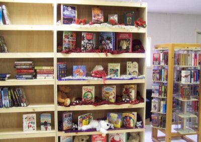 A set of wooden bookshelves is decorated with Christmas books, tinsel, and small holiday displays, next to a glass display case filled with additional titles. The shelves contain a mix of seasonal items and general reading materials.