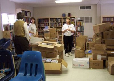 Several people stand inside a temporary library area surrounded by open cardboard boxes filled with books, with full bookshelves lining the back wall. Additional boxes and scattered supplies cover the floor.