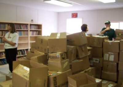 A group of people stands among tall stacks of unpacked cardboard boxes inside a temporary library space, with bookshelves lining the walls. Bright overhead lighting illuminates the room as materials are being sorted and organized.
