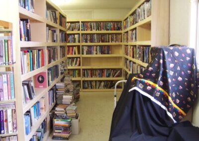 Rows of wooden bookshelves filled with books create a narrow aisle in the temporary Pass Christian Library, with stacks of additional books placed on the floor. A draped cart sits near the window at the end of the aisle.