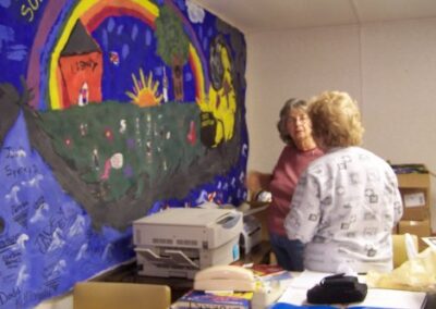 Two people stand at a table inside the temporary Pass Christian Library, with a large colorful mural featuring a rainbow, sun, and landscape filling the wall behind them. The desk is covered with office supplies, papers, and equipment.