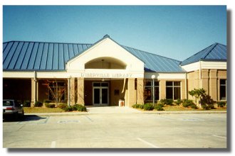 Single‑story library building with a blue metal roof, arched entrance, and large front windows, with a parking area in the foreground.