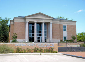 Exterior view of a two‑story brick library building with tall white columns at the entrance, large vertical windows, and a sign in front that reads “Harrison County Library System.”
