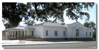A white, single‑story public building with large windows, viewed from across the street under tree branches.
