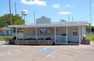 A small white building with a front porch and railing, shown from a parking lot with marked spaces.