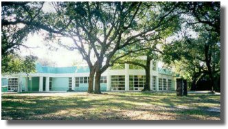 Blue colored building with large windows, partially shaded by tall trees in the foreground.
